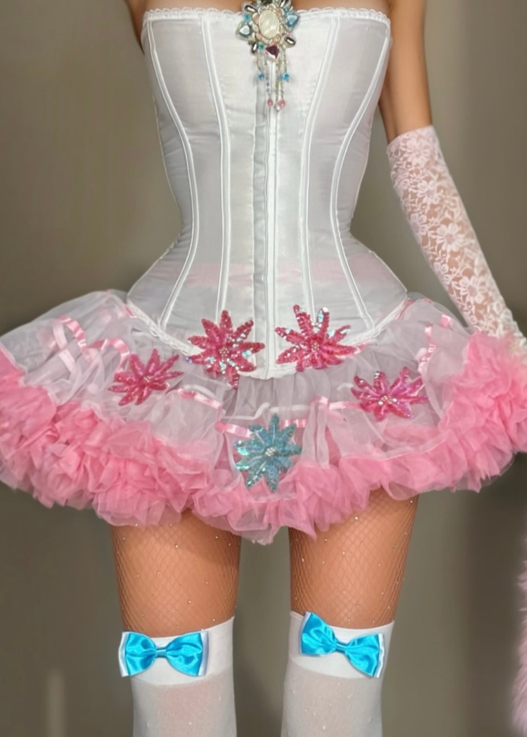 A close-up of a doll or mannequin torso wearing a white corset-style dress with a pink ruffled tutu, pink and blue snowflake embellishments, white fishnet stockings, and lace arm sleeves.