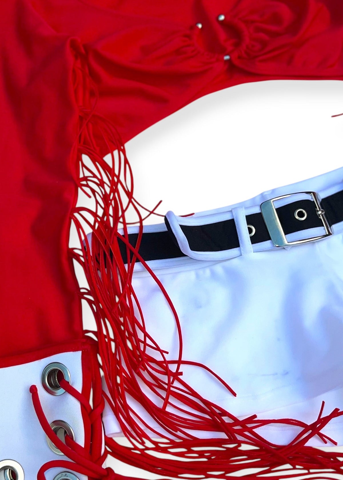 A close-up of a costume or outfit featuring a bright red fringed top and matching red fringe trim over white bottoms, secured with a black belt and silver buckle.