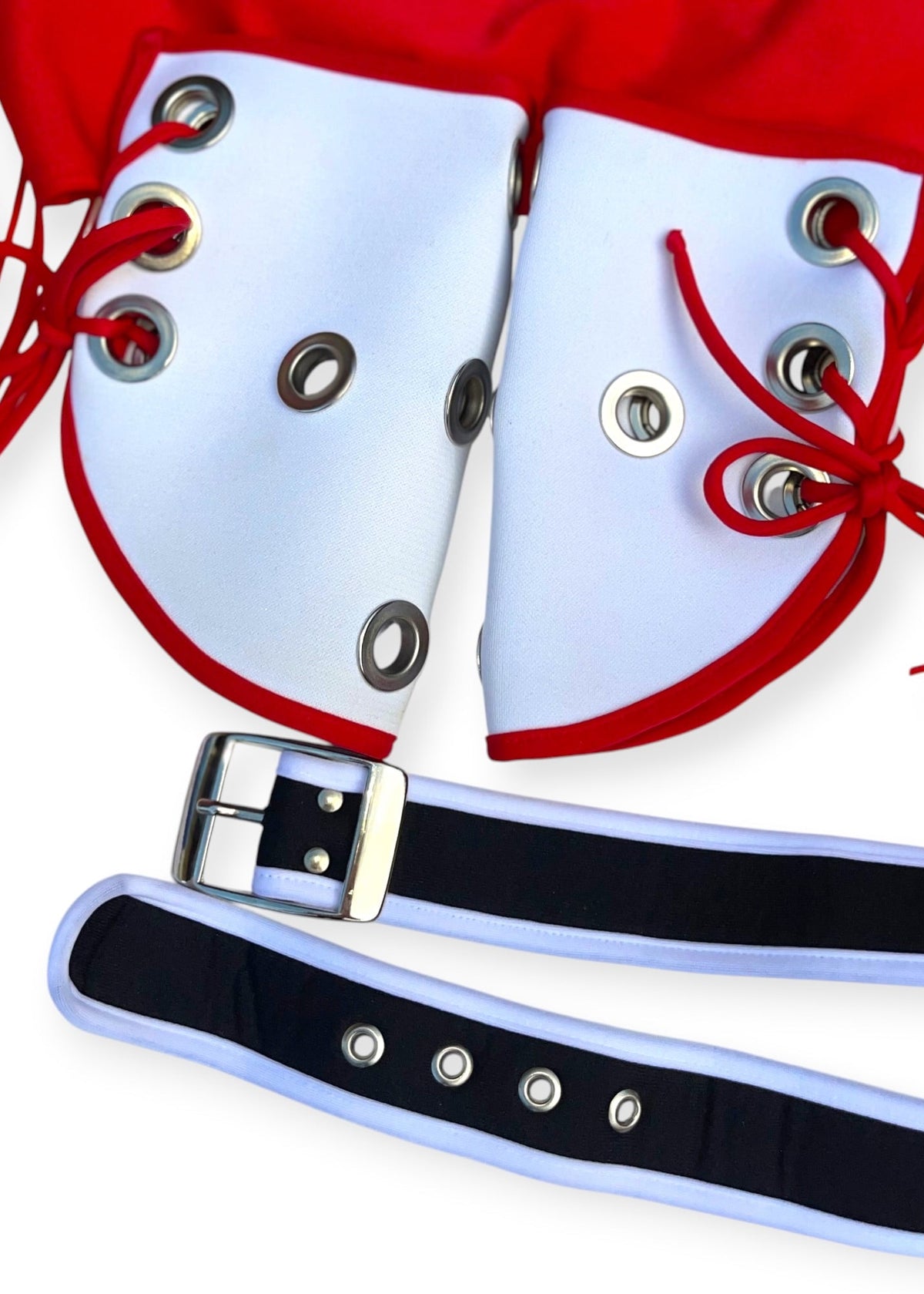 A close-up of a costume accessory set featuring two white padded shoulder pieces with red trim and red laces, a red fabric backing with fringe, a black and white adjustable strap with a silver buckle, and silver metal grommets.