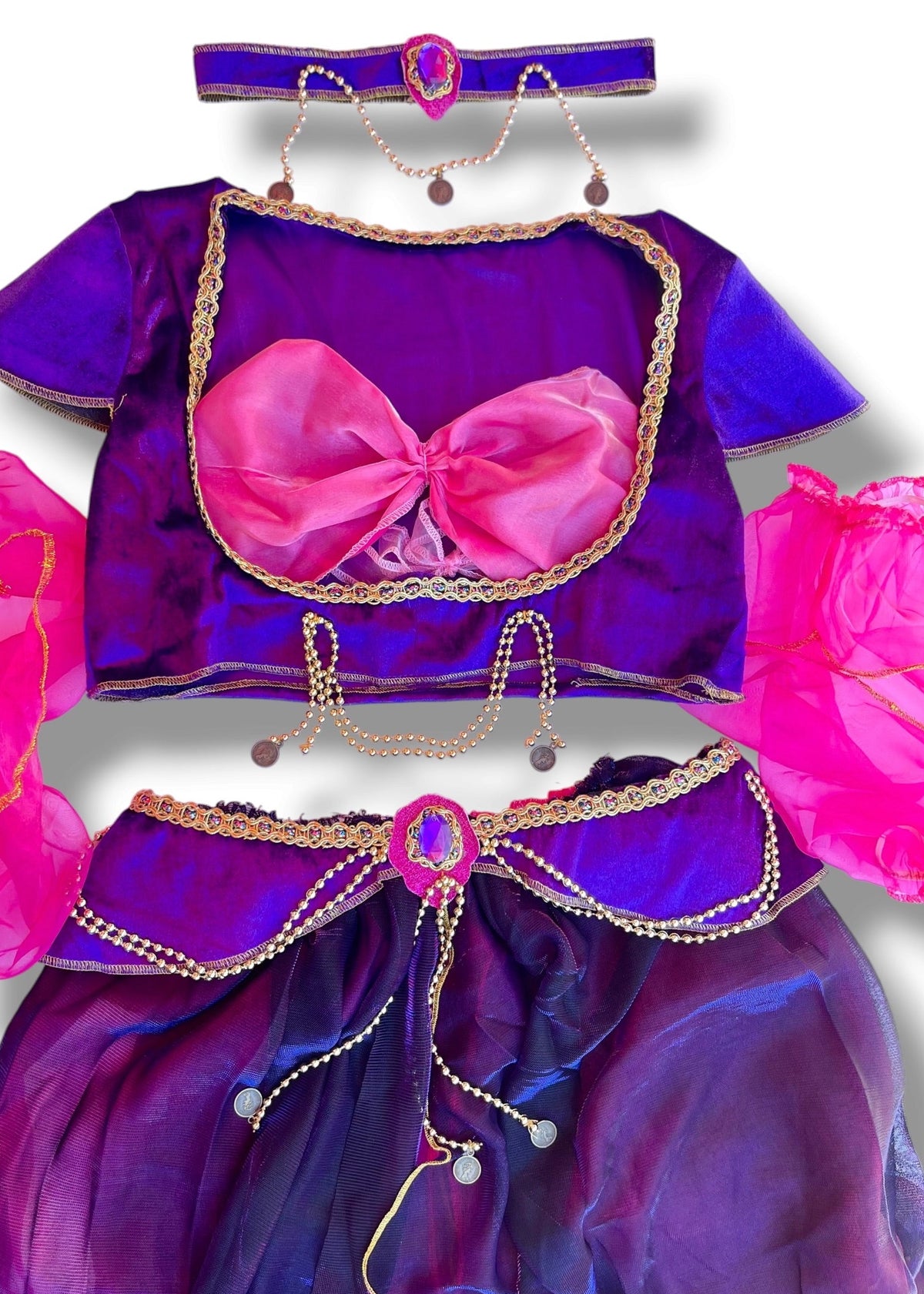 A children's genie-style costume set in purple and pink, featuring a short-sleeve top with a large pink bow, a matching skirt with gold trim and drawstring, bright pink arm pieces, and a headband with dangling charms, shown on a white background.