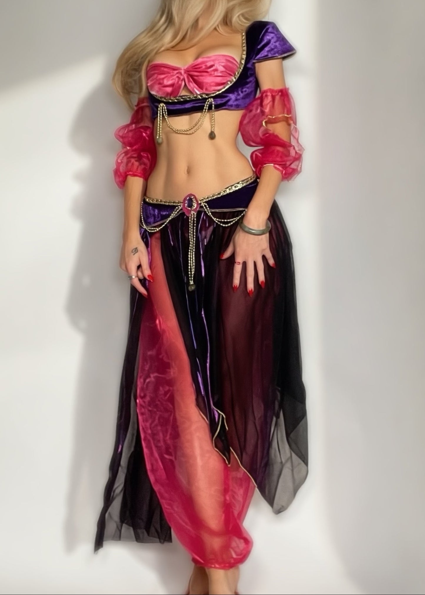 A woman modeling a genie-style costume with a shiny purple and pink bra top, puffed pink sleeves, a dark purple and pink skirt, and gold chain accents, posed against a plain light gray wall.