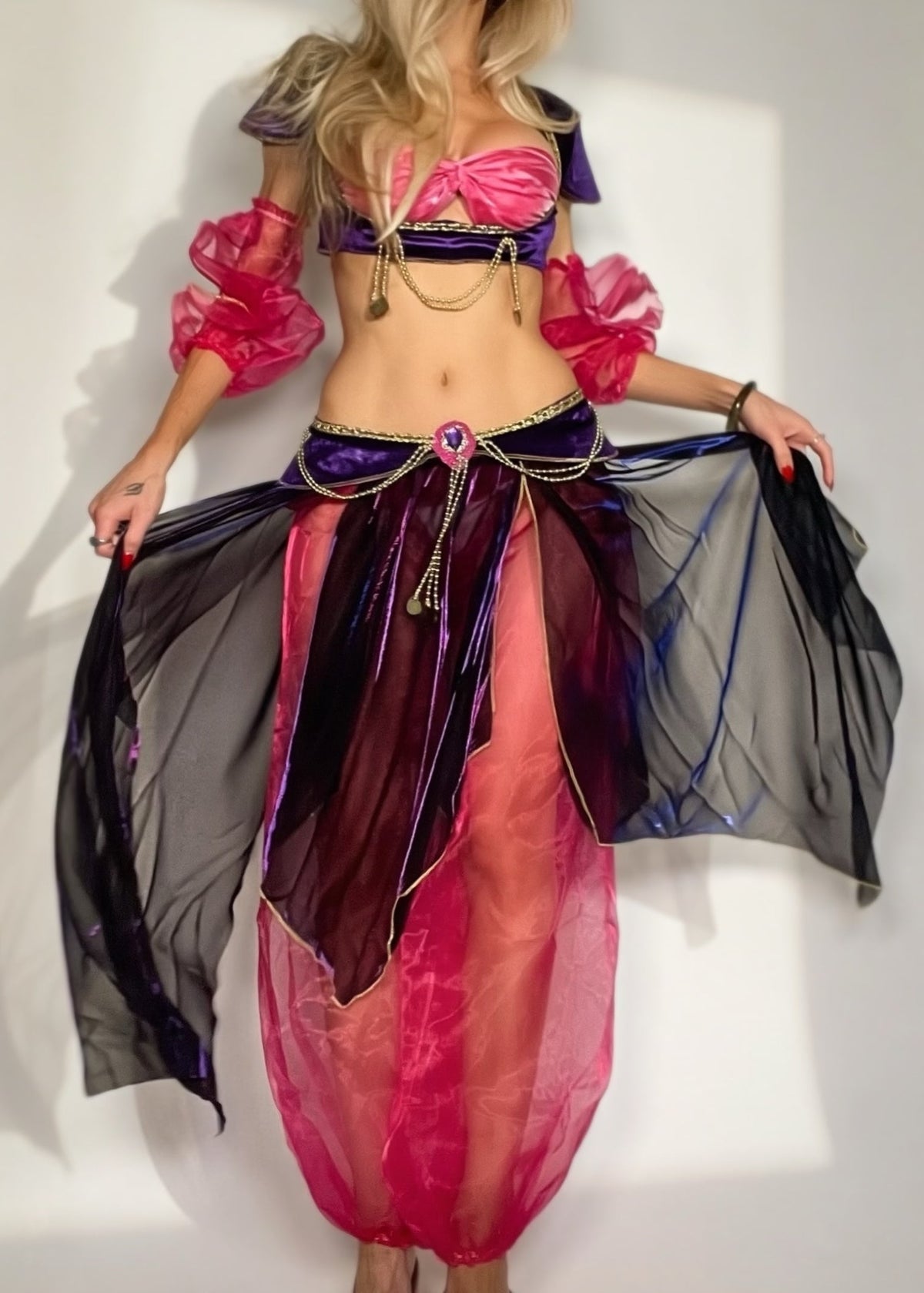 A woman modeling a belly dance costume set consisting of a metallic purple bra top, bright pink ruffled sleeves, a purple waist scarf with gold chain details, and sheer black harem pants with a pink and purple gradient.