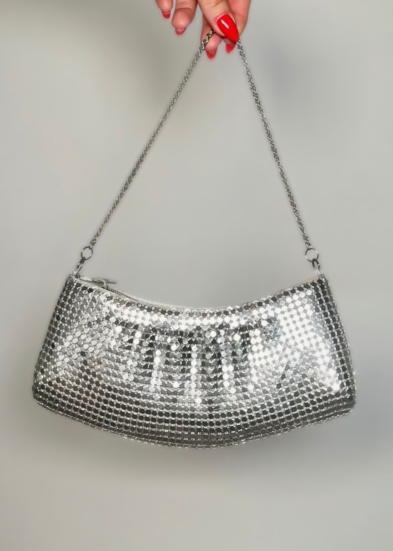Silver mesh handbag held by a hand with red nail polish against a gray background